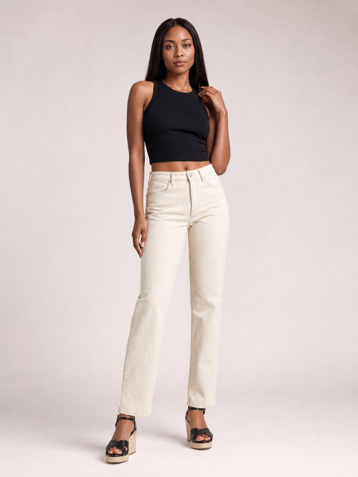 A model poses in a fitted black cutout round-neck tank with sleeveless styling and a cropped silhouette, paired with high-waisted cream jeans.
