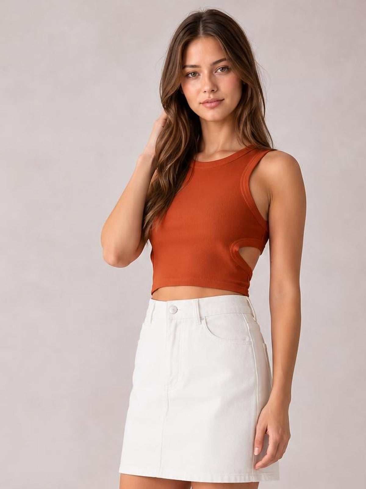 Model poses in a rust-orange cutout round neck tank with wide straps, a fitted cropped silhouette, and a side waist opening.