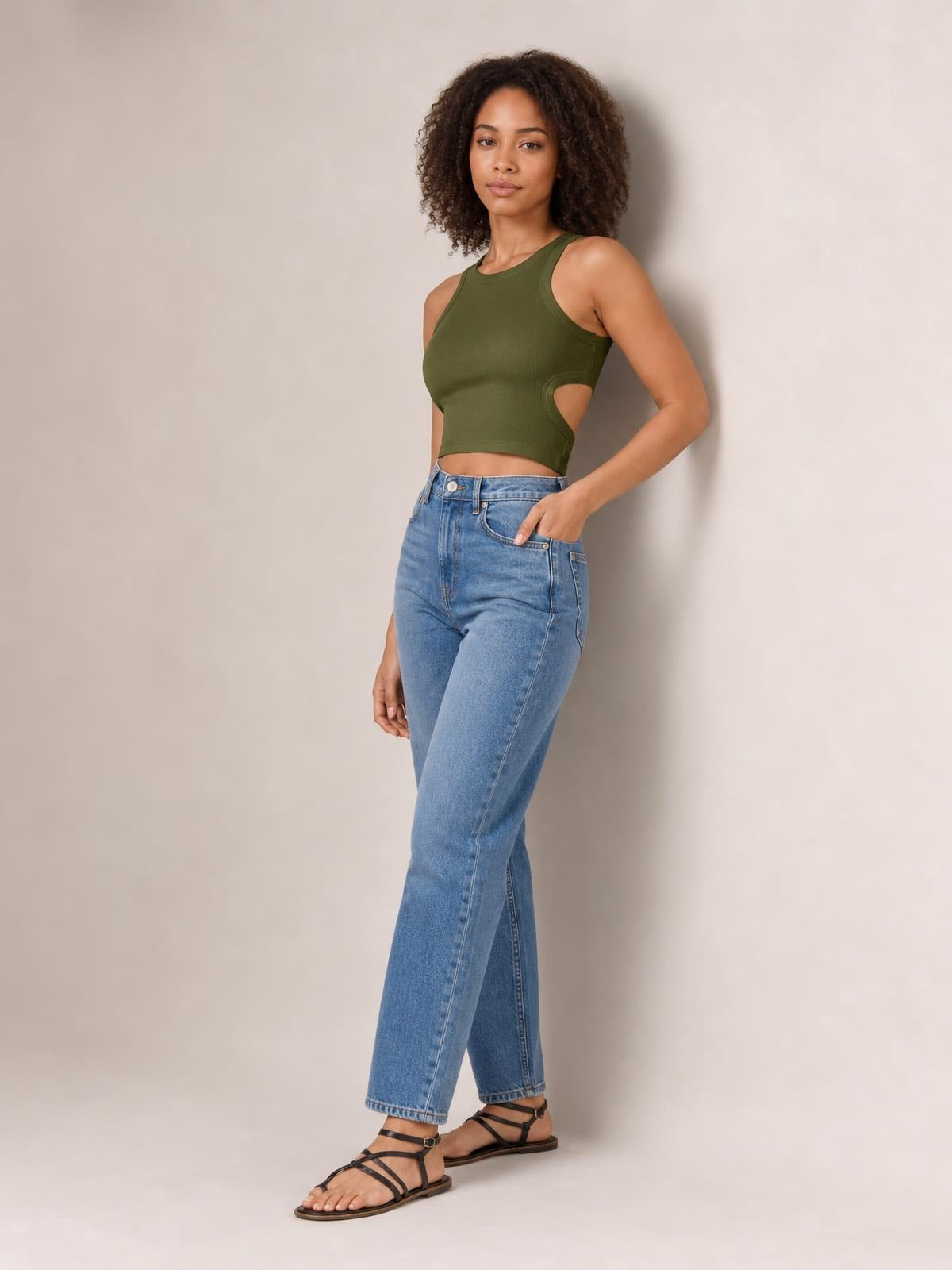 A model poses in an olive green cutout round neck tank with a fitted cropped silhouette, wide shoulder straps, and side cutout detailing.