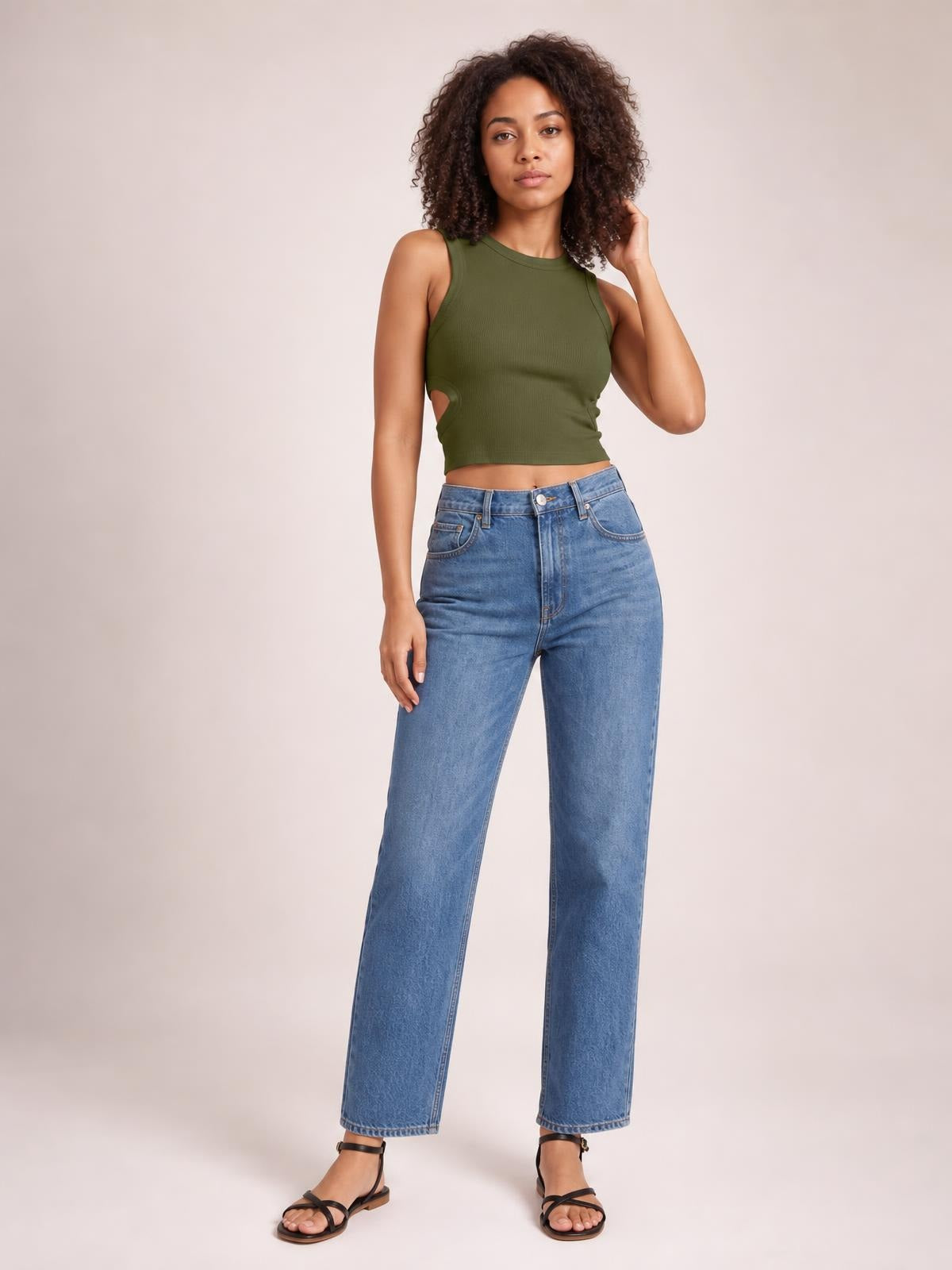 Model wearing an olive green cutout round neck tank with a fitted cropped silhouette, standing in blue straight-leg jeans with one hand touching her hair.