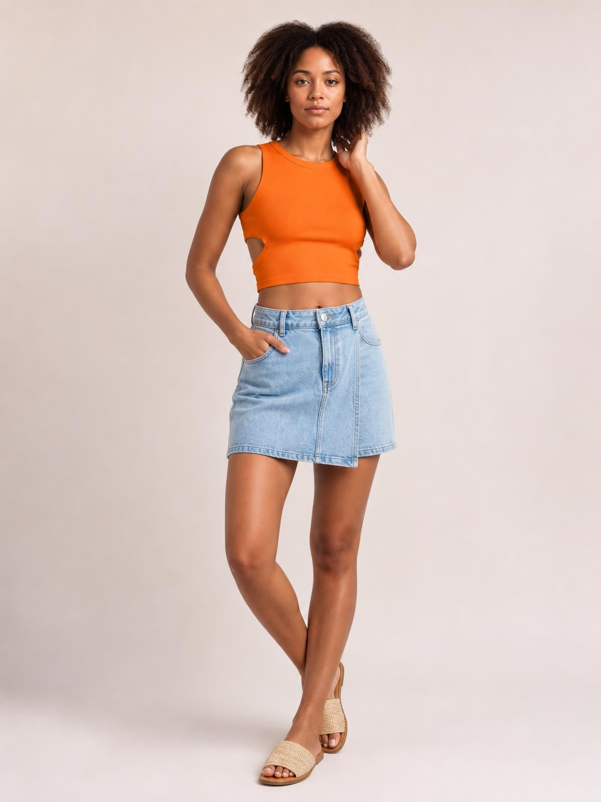 A model poses in an orange cutout round neck tank with a fitted cropped silhouette, wide shoulder straps, and her legs crossed.