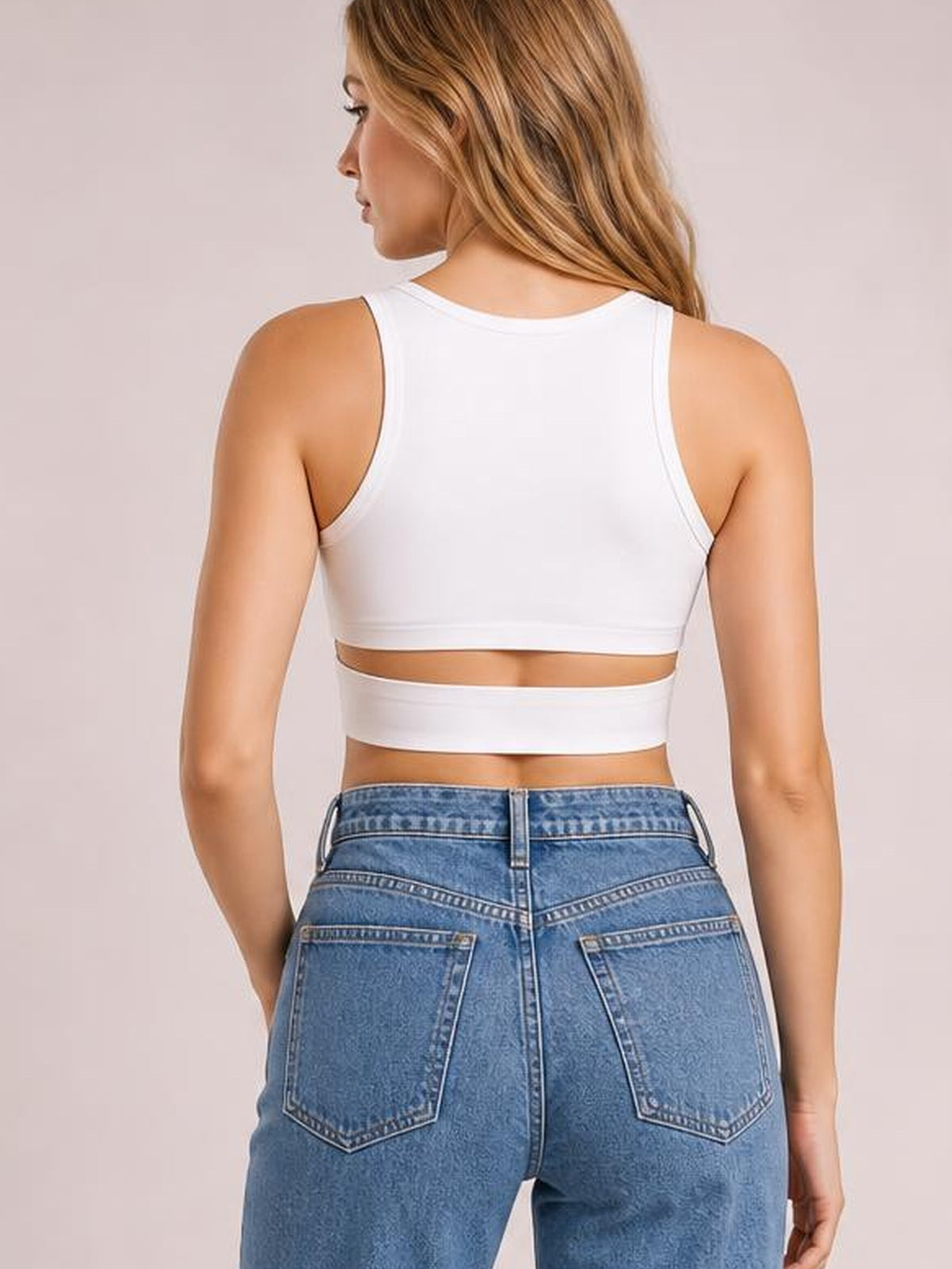 A model poses in profile wearing a white cutout round neck tank with a cropped, fitted silhouette, wide shoulder straps, and an open back detail above blue jeans.