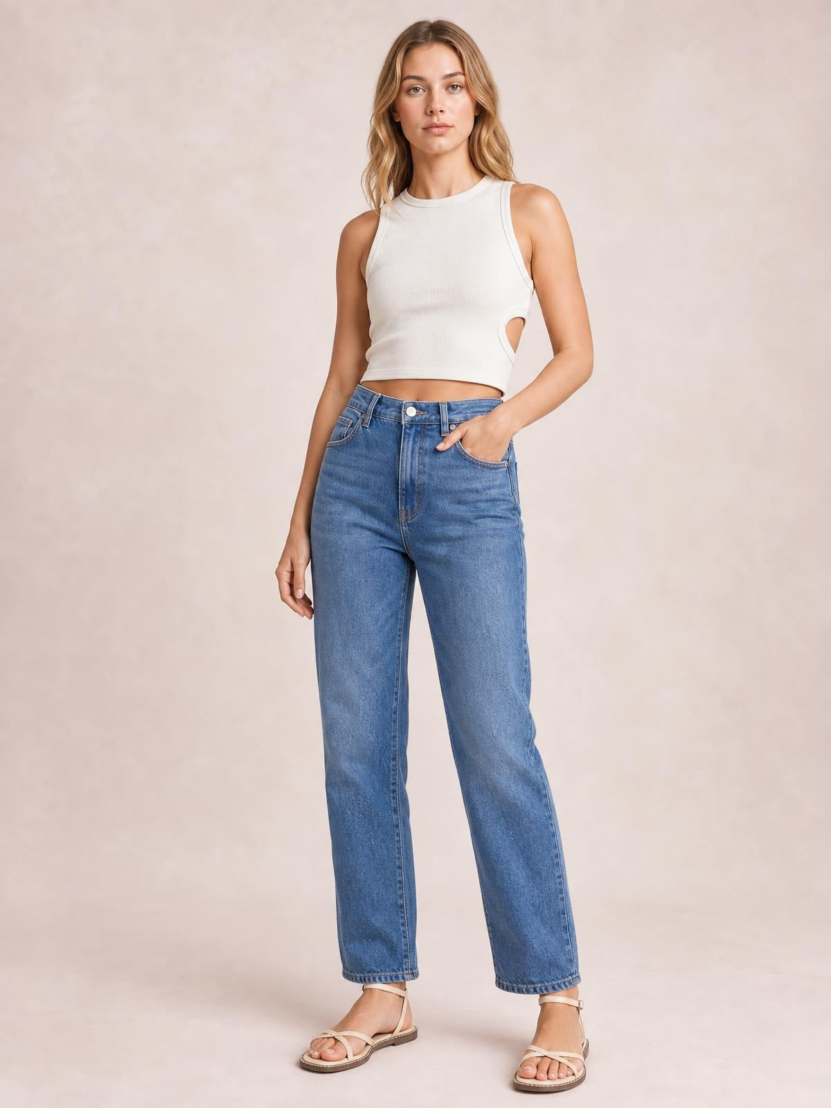 Model wears a white cutout round neck tank with a fitted cropped silhouette and wide shoulder straps, posing with one hand in her jeans pocket.