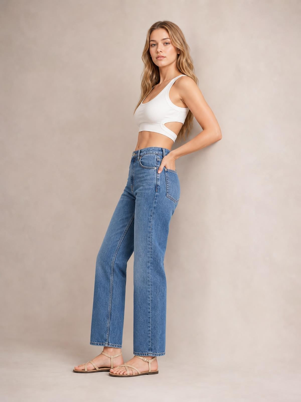 Model wears a white cutout round neck tank with a fitted cropped silhouette and side opening, standing in a relaxed pose with one hand in her jeans pocket.