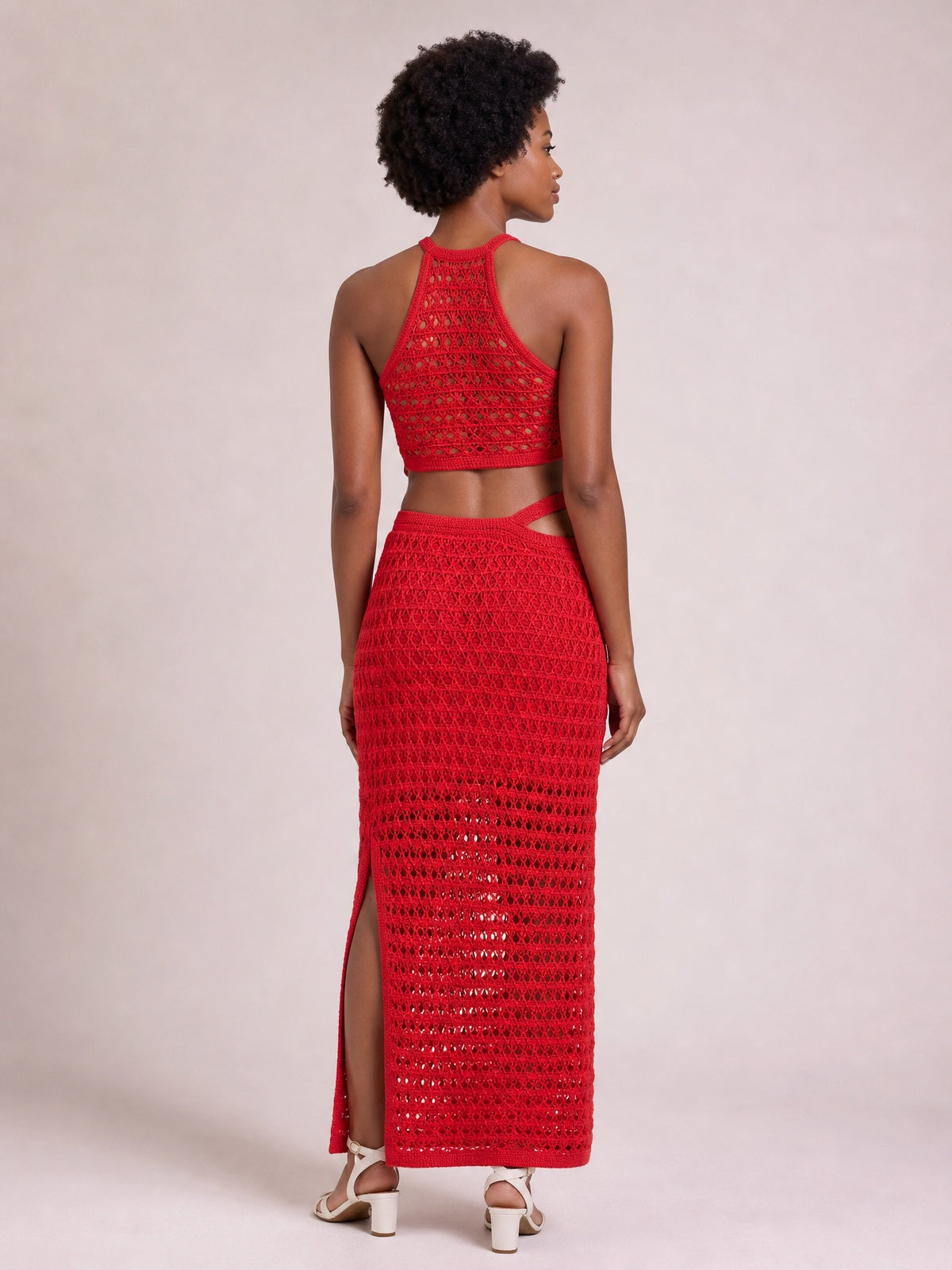 A model stands facing away in a red open-knit sleeveless sweater dress with a fitted midi silhouette, waist cutout, and side slit.