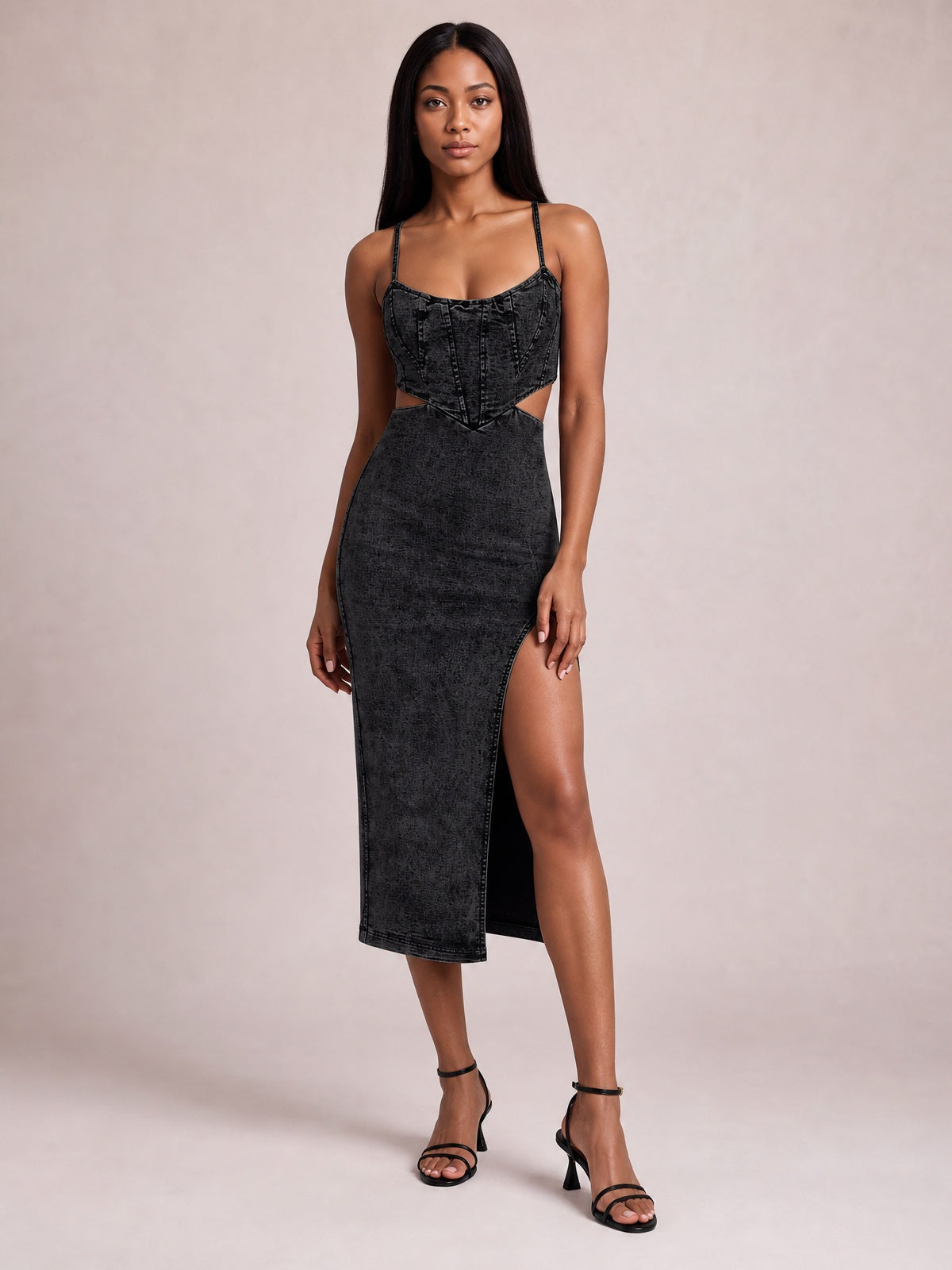A model stands in a black denim midi dress with spaghetti straps, a fitted seamed bodice, waist cutouts, and a high side slit creating a body-skimming silhouette.