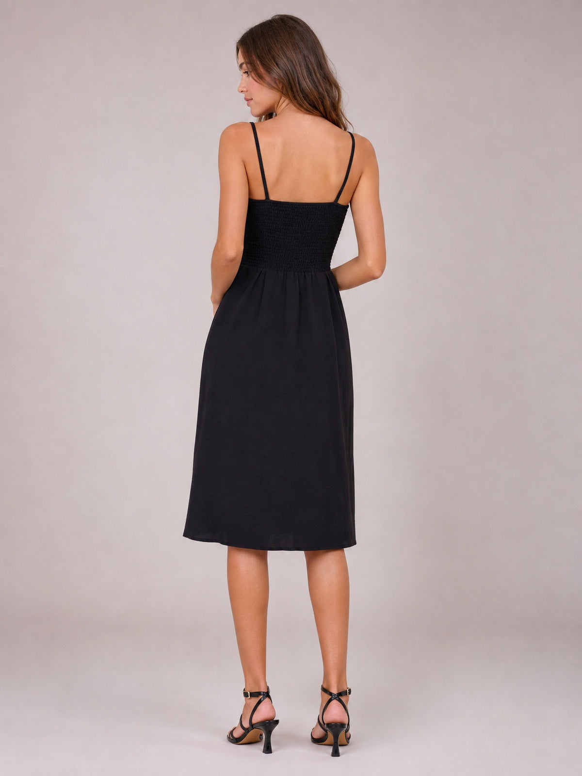 A model stands turned away in a black smocked cami dress with slender shoulder straps, a fitted bodice, and a softly flowing below-the-knee A-line skirt.
