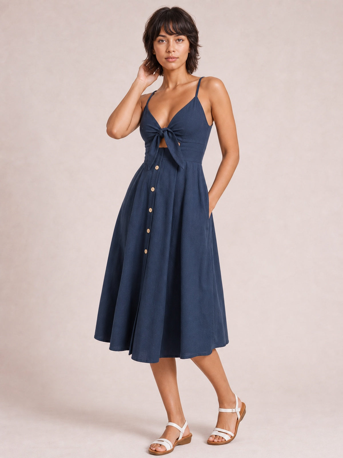 Model wearing a navy cami dress with slender straps, sweetheart neckline, tied cutout bodice, decorative front buttons, and a flowing below-knee A-line skirt.