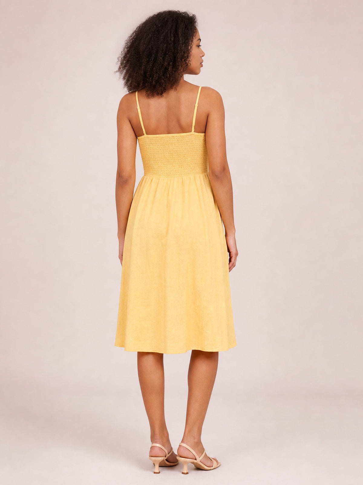A model stands turned away in a sunny yellow cami dress with slender straps, a smocked back bodice, and a gently full below-the-knee A-line skirt.