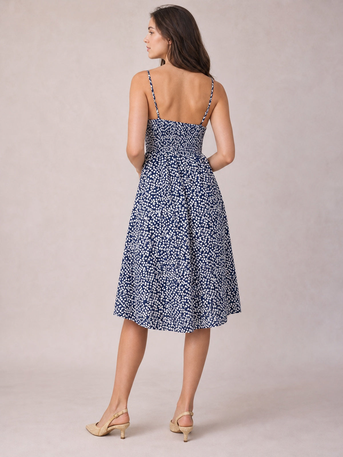 Model poses turned away in a navy floral cami dress with slender straps, a smocked back, fitted waist, and gently flared below-the-knee A-line skirt.