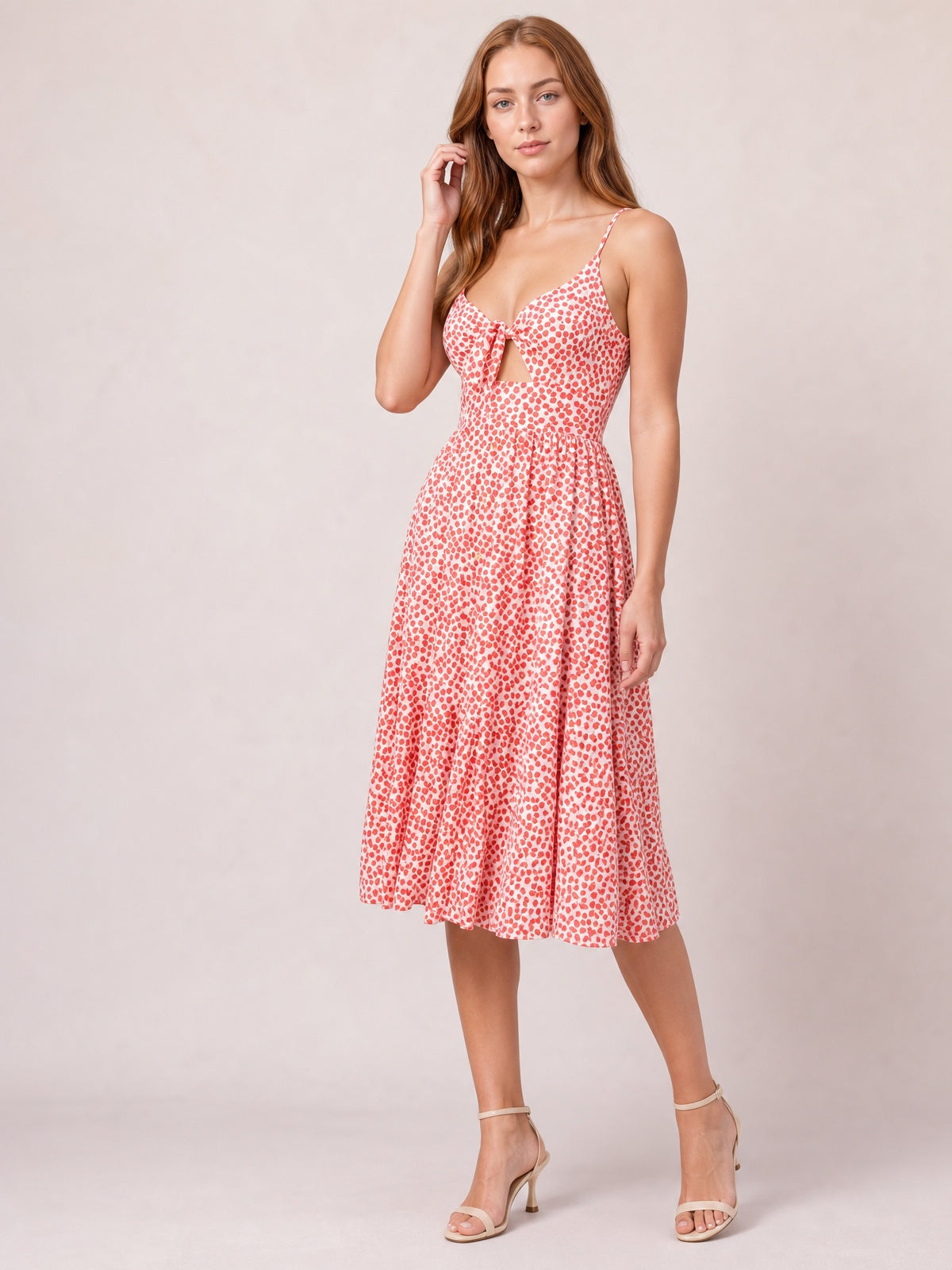 Model poses in a coral-and-white spotted cami dress with slender straps, a tied sweetheart cutout bodice, fitted waist, and softly full below-the-knee A-line skirt.