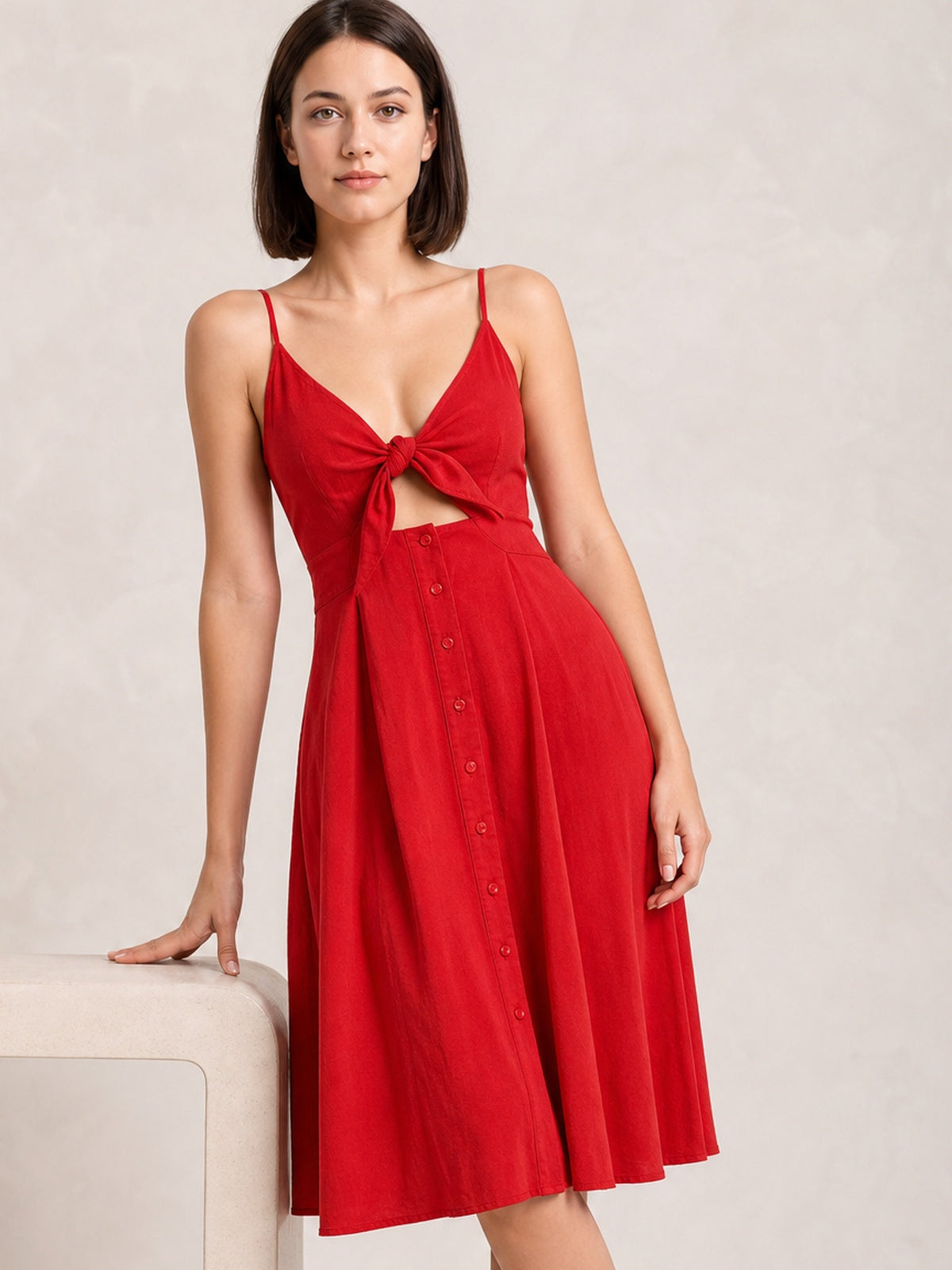 A model poses in a red cami dress with slender straps, a tied sweetheart neckline and front cutout, decorative buttons, and a flowing A-line skirt.