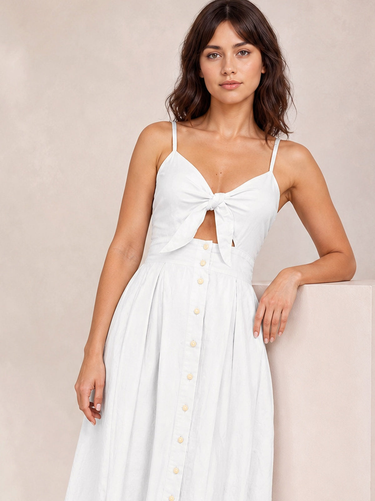 Model wears a white cami dress with slender straps, a tied sweetheart cutout bodice, front decorative buttons, and a softly gathered A-line skirt.