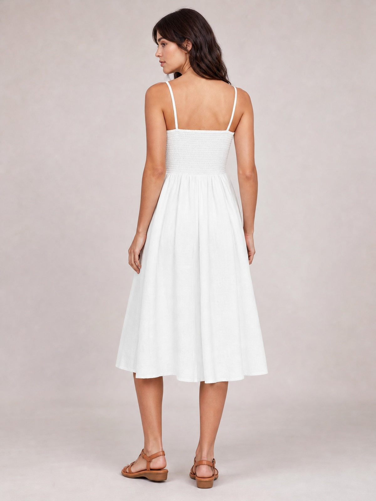 Model shown from the back wearing a white cami dress with slender straps, smocked bodice, gathered waist, and flowing below-the-knee A-line skirt.
