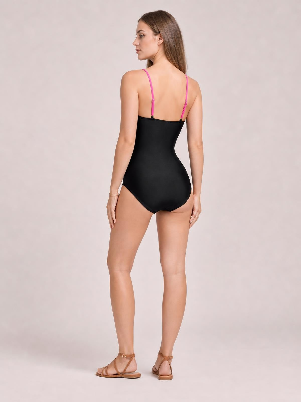 Model poses with her back turned in a black cutout spaghetti strap one-piece swimsuit, featuring a sleek fitted silhouette and contrasting pink straps.