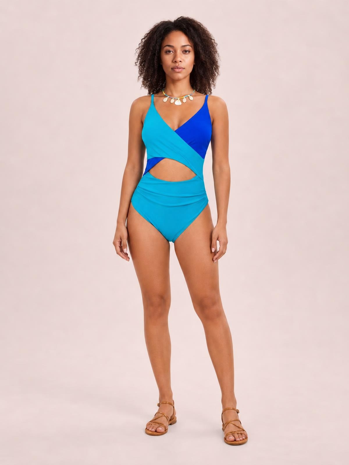 Model stands in a two-tone turquoise and royal blue one-piece swimwear with spaghetti straps, a wrap-style bodice, waist cutout, and fitted high-rise silhouette.