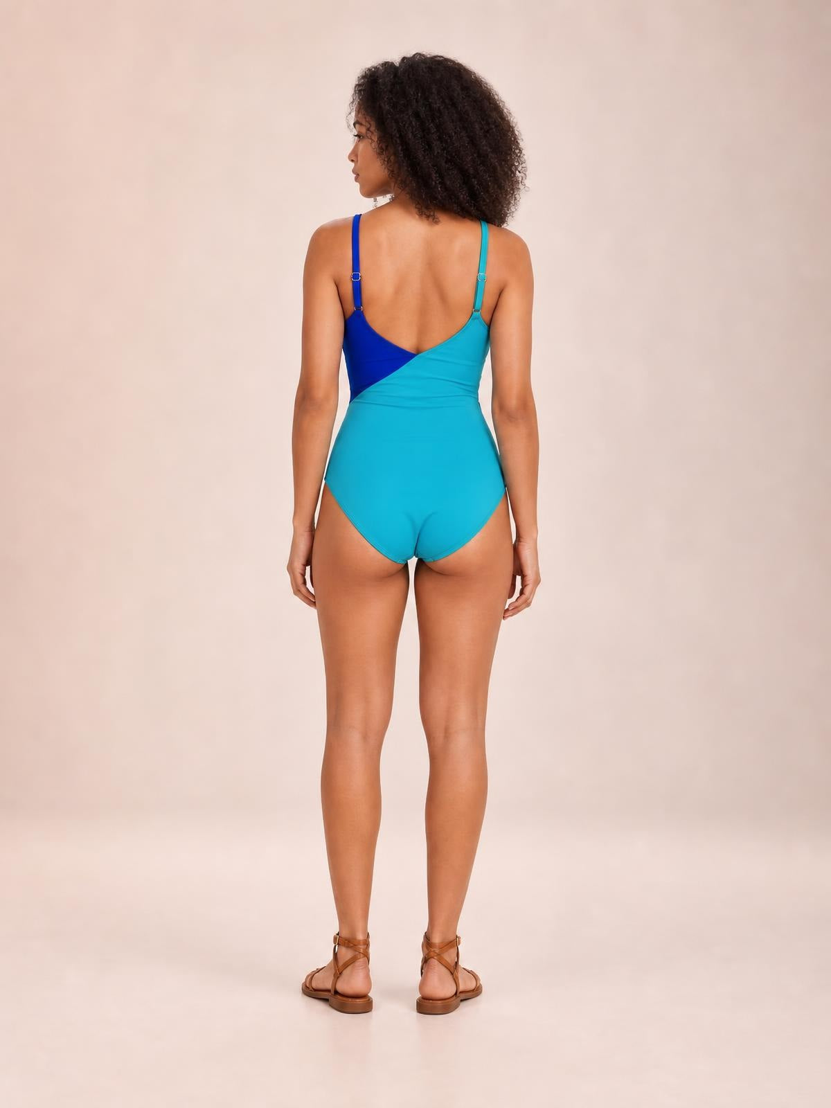 Model stands facing away in a turquoise and royal blue color-block cutout one-piece swimsuit with spaghetti straps, a sculpted silhouette, and high-cut legs.