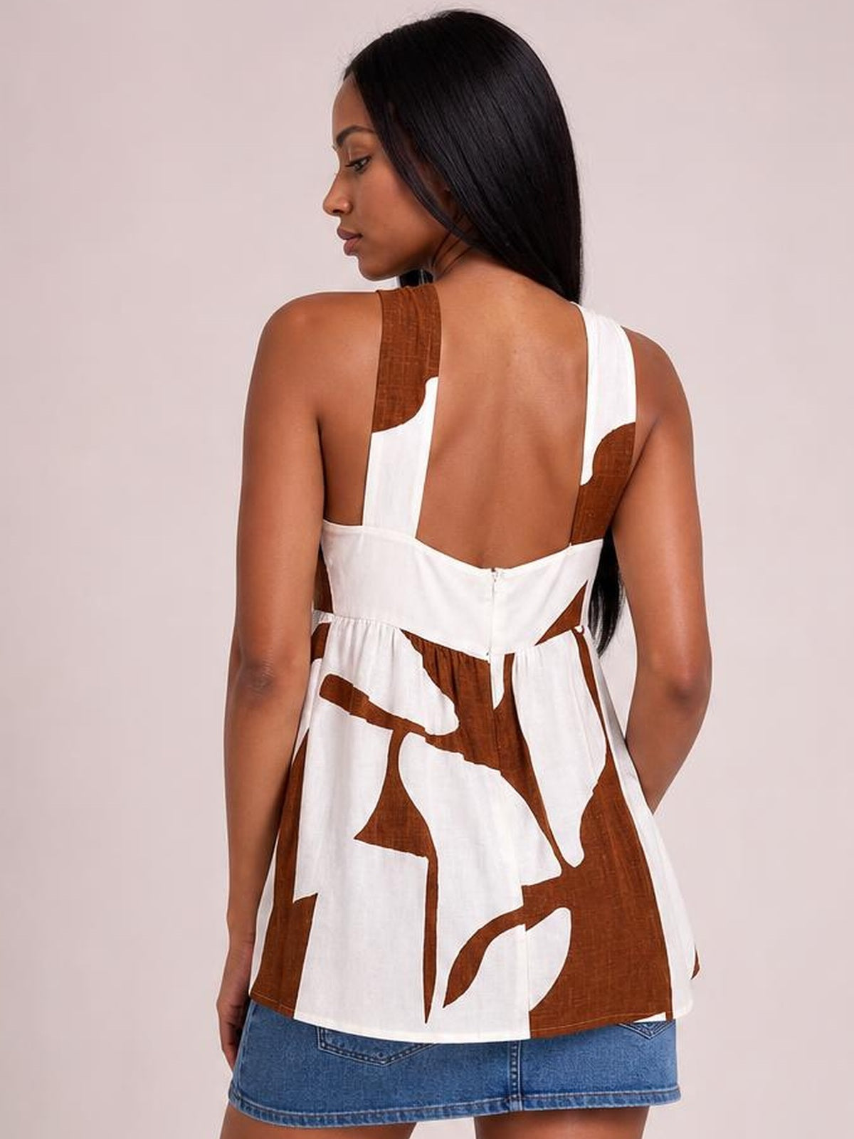 Model turned away wears a white and brown abstract-print cami with wide straps, an open back, and a ruched waist creating a relaxed peplum silhouette.