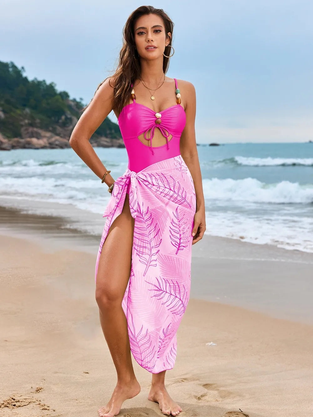 Model stands with one hand on her hip in a bright pink cutout sweetheart-neck one-piece swimsuit with ruched bust and a pink leaf-print cover-up.
