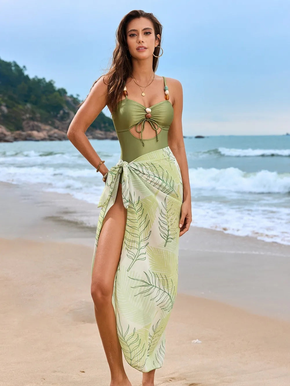 Model poses in an olive-green cutout sweetheart-neck one-piece swimsuit with gathered bust and tie detail, paired with a leafy wrap cover-up.