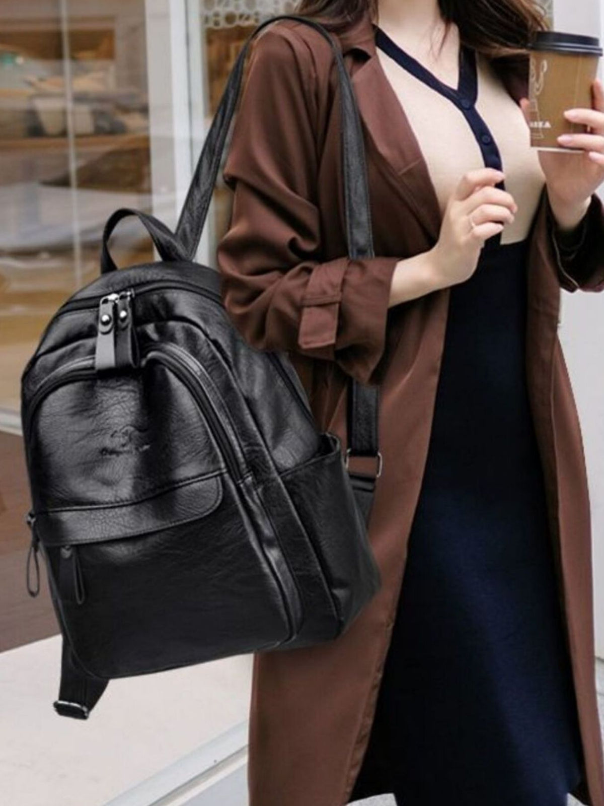 A woman carries a medium black PU leather backpack with adjustable straps, a rounded silhouette, zippered compartments, and a front pocket.