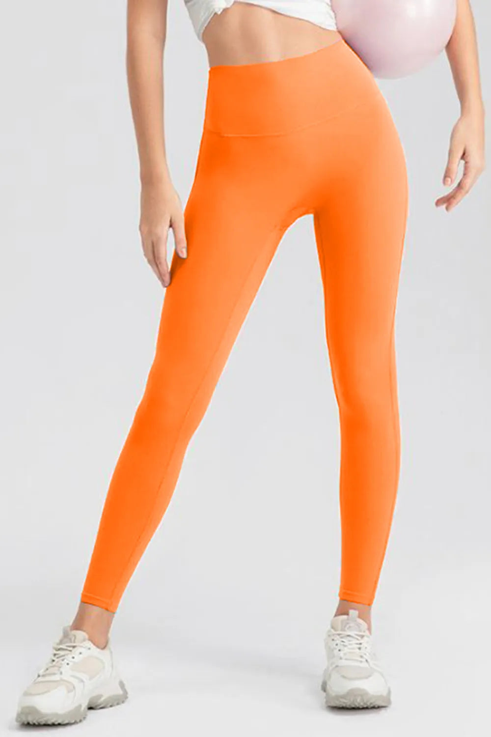 Model wearing bright orange high-waist skinny active leggings with a smooth wide waistband and full-length fitted silhouette, standing with legs apart.