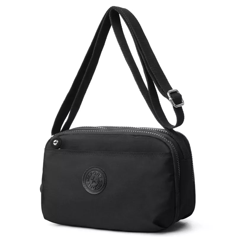 Small black nylon crossbody bag with an adjustable strap, rounded rectangular silhouette, dual zip compartments, and a front embossed logo.
