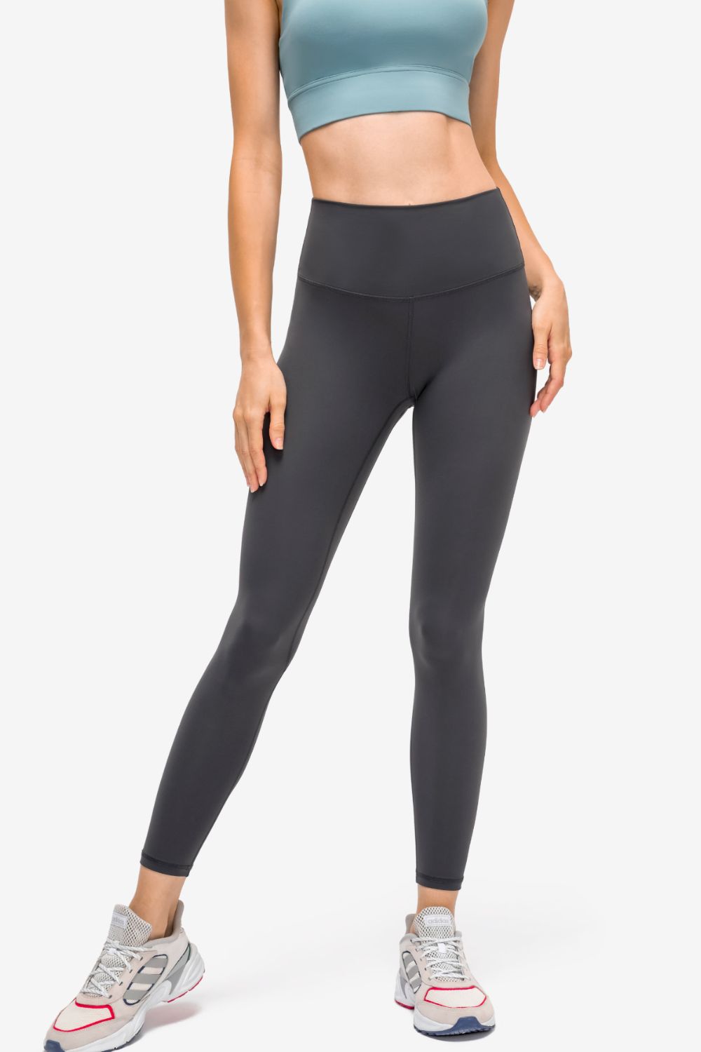 Model stands with one leg angled in dark gray high-rise sports leggings, showing a slim fit, matte fabric, curved waistband seam, and tapered ankle construction.