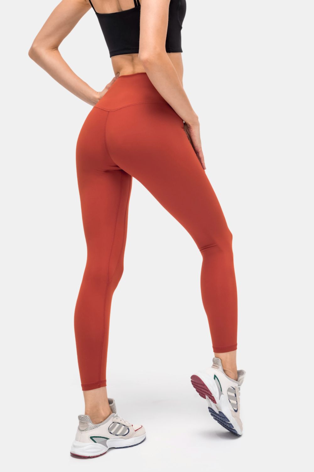 A model poses from behind in solid rust-red high-rise sports leggings with a slim silhouette, matte fabric, and curved back seam construction.