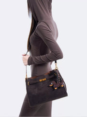 A model wears a medium dark brown suede crossbody bag with a structured rectangular silhouette, decorative buckle closure, and gold-tone hardware.