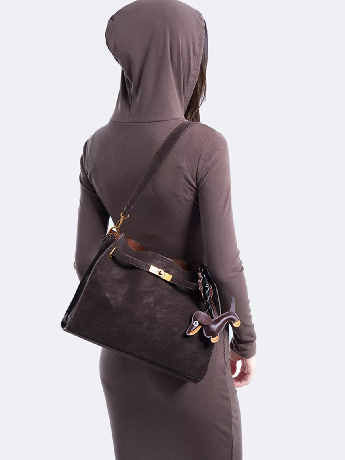 A model shown from behind wears a medium dark brown suede decorative buckle crossbody bag with a structured silhouette, long strap, and gold-tone hardware.