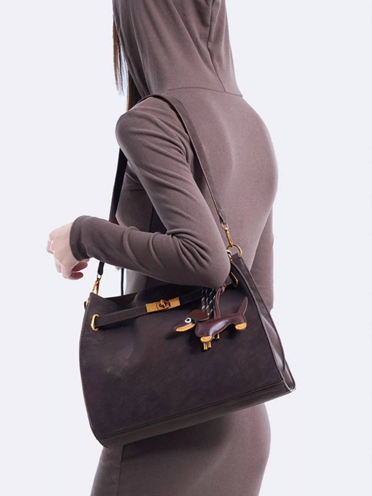 Model wears a medium dark brown suede crossbody bag with a structured rectangular silhouette, decorative gold buckle, top handle, and long adjustable strap.