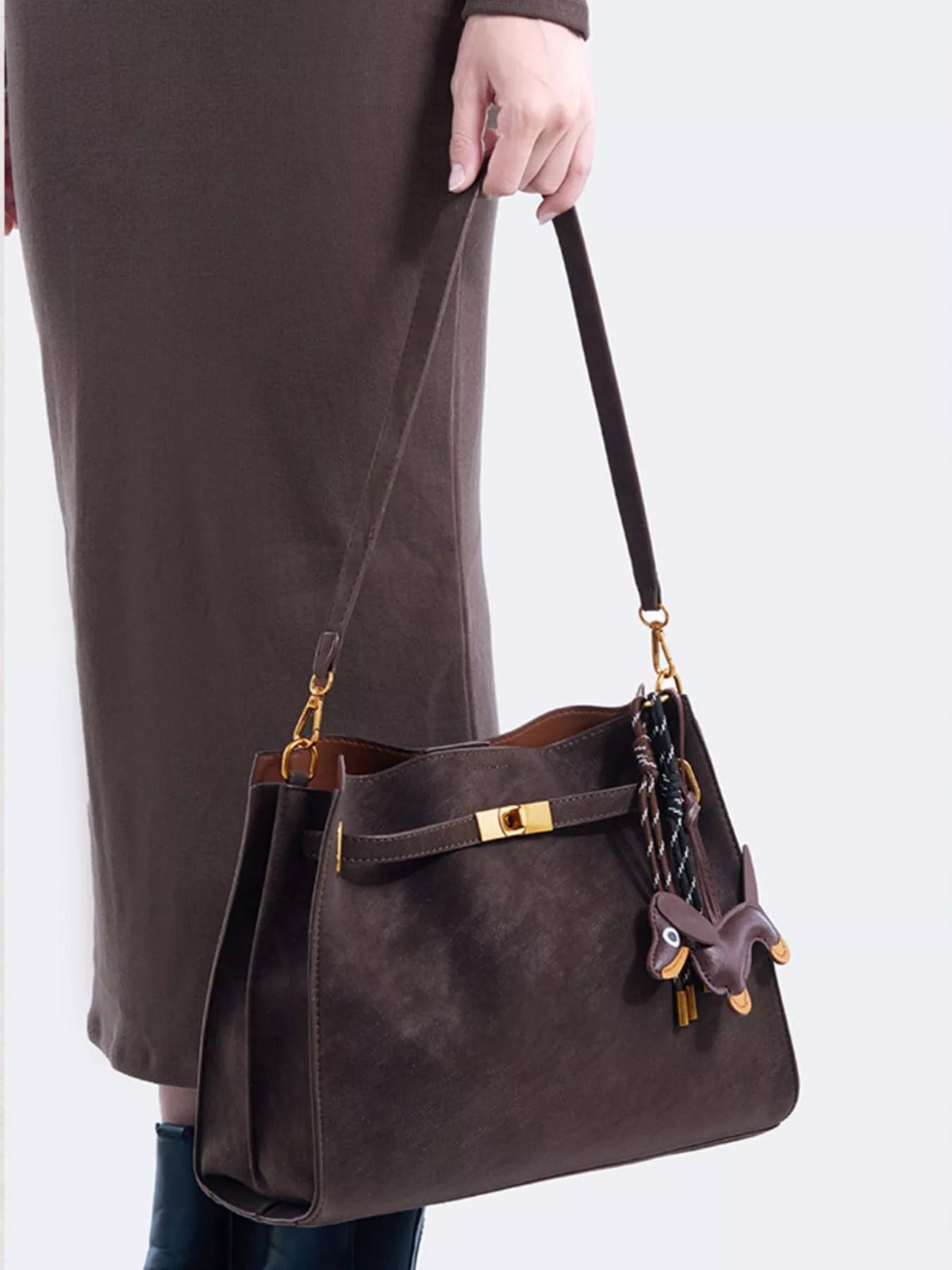 A model holds a medium dark brown suede crossbody bag with a structured silhouette, decorative gold buckle, long strap, and playful hanging charm.