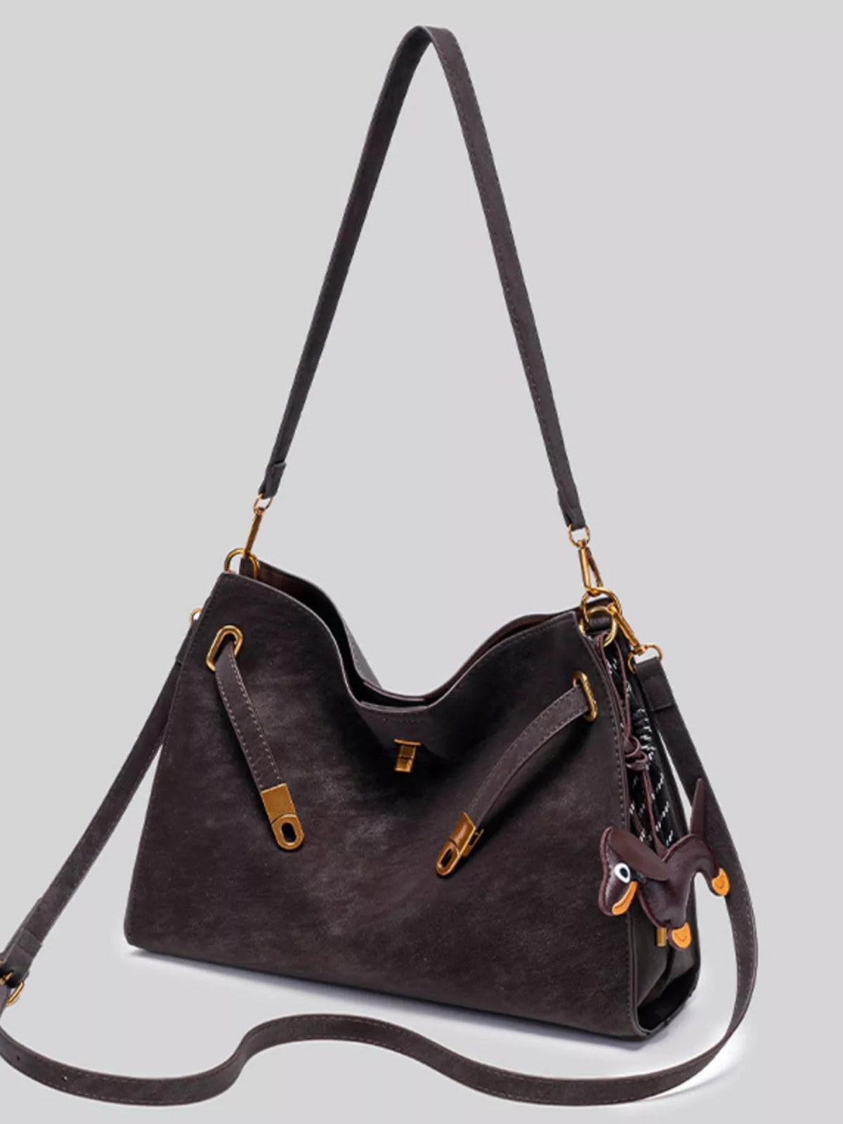 Medium dark brown suede crossbody bag with a structured satchel silhouette, decorative buckle straps, gold-tone hardware, and a long adjustable shoulder strap.
