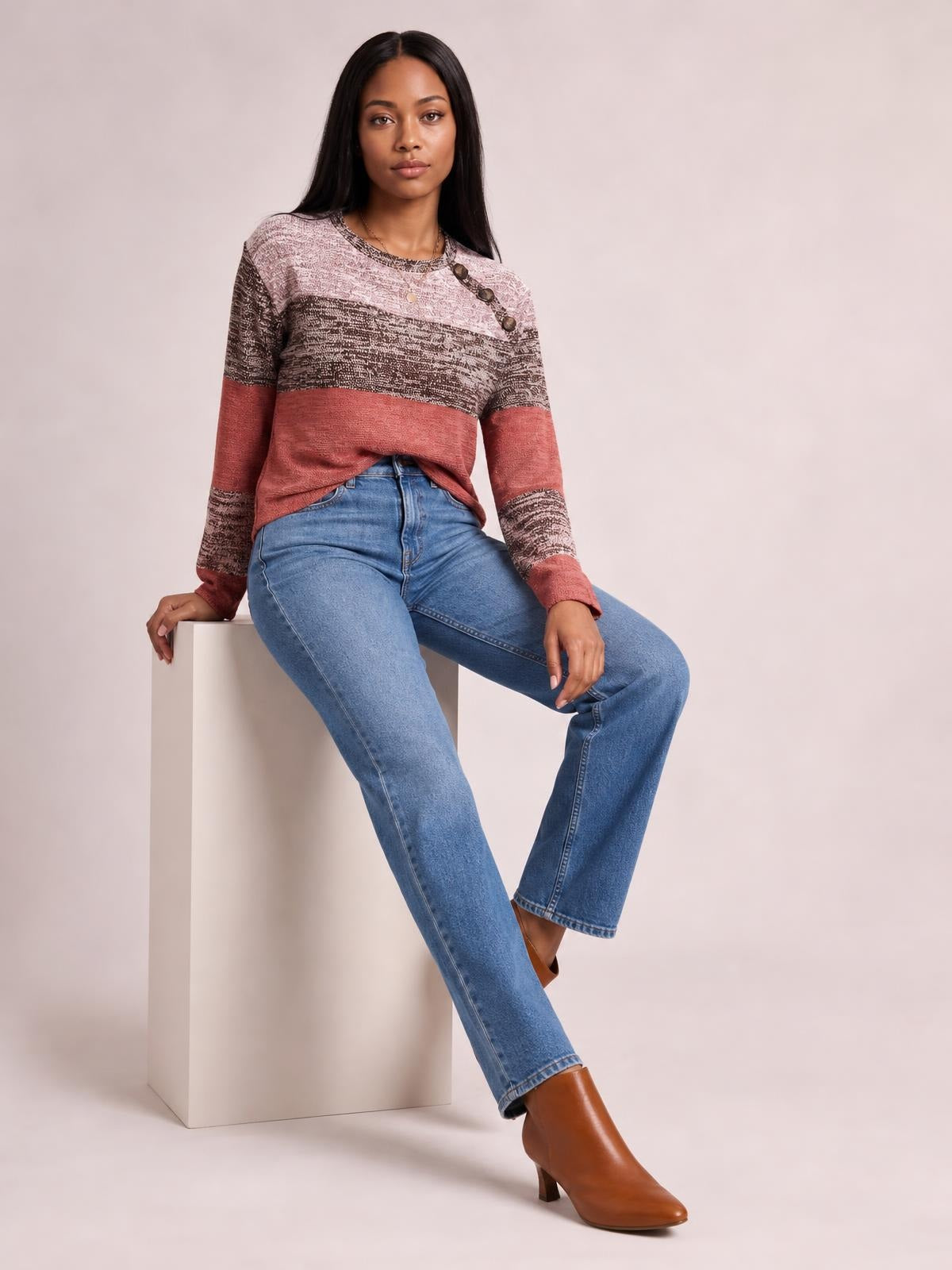 A seated model wears a color-blocked long-sleeve T-shirt with a contrast round neck, decorative buttons at the shoulder, and a relaxed silhouette.