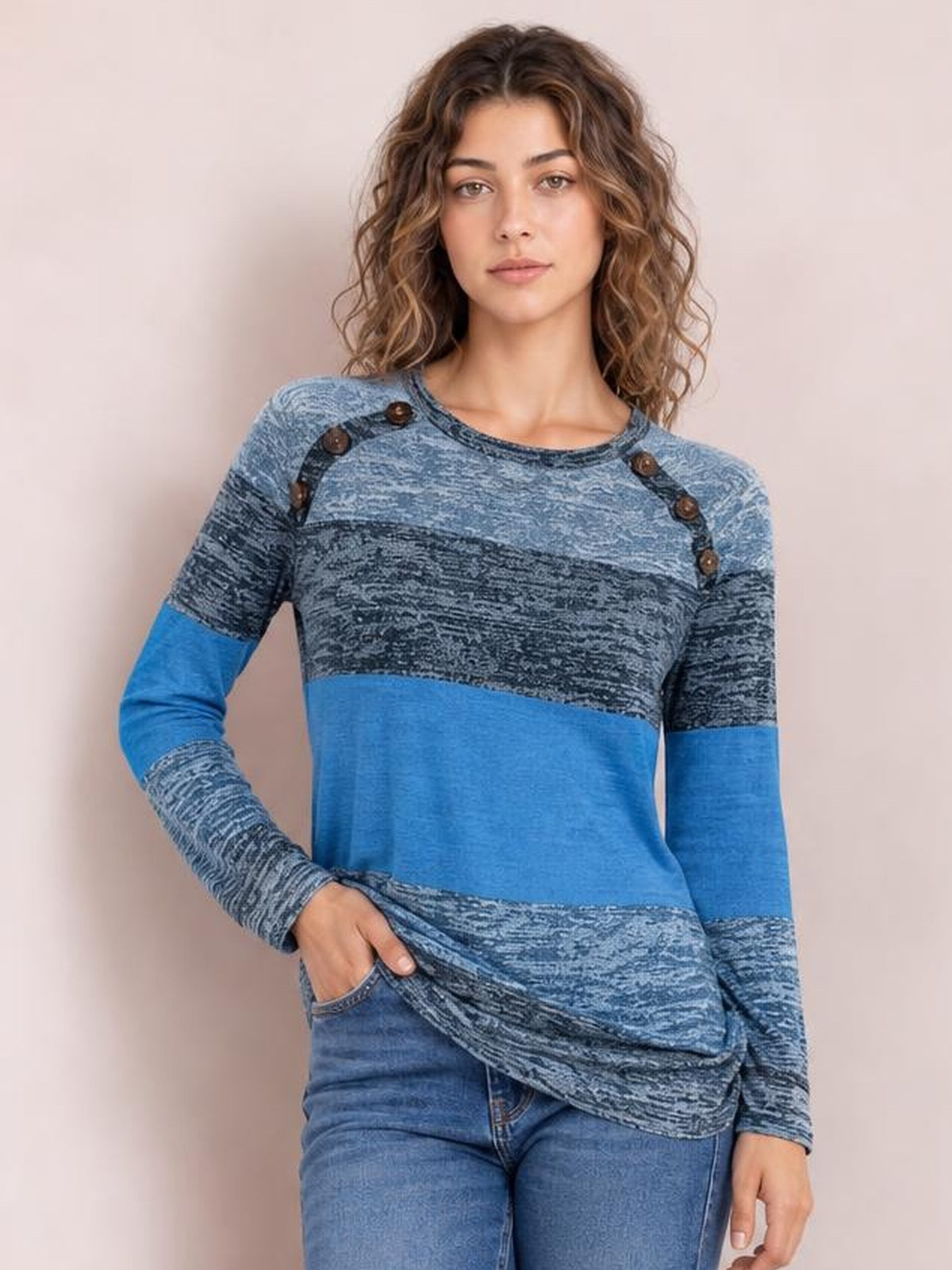 Model wears a blue contrast long-sleeve T-shirt with a round neck, decorative button accents at the shoulders, and a relaxed silhouette, posing with one hand in her pocket.