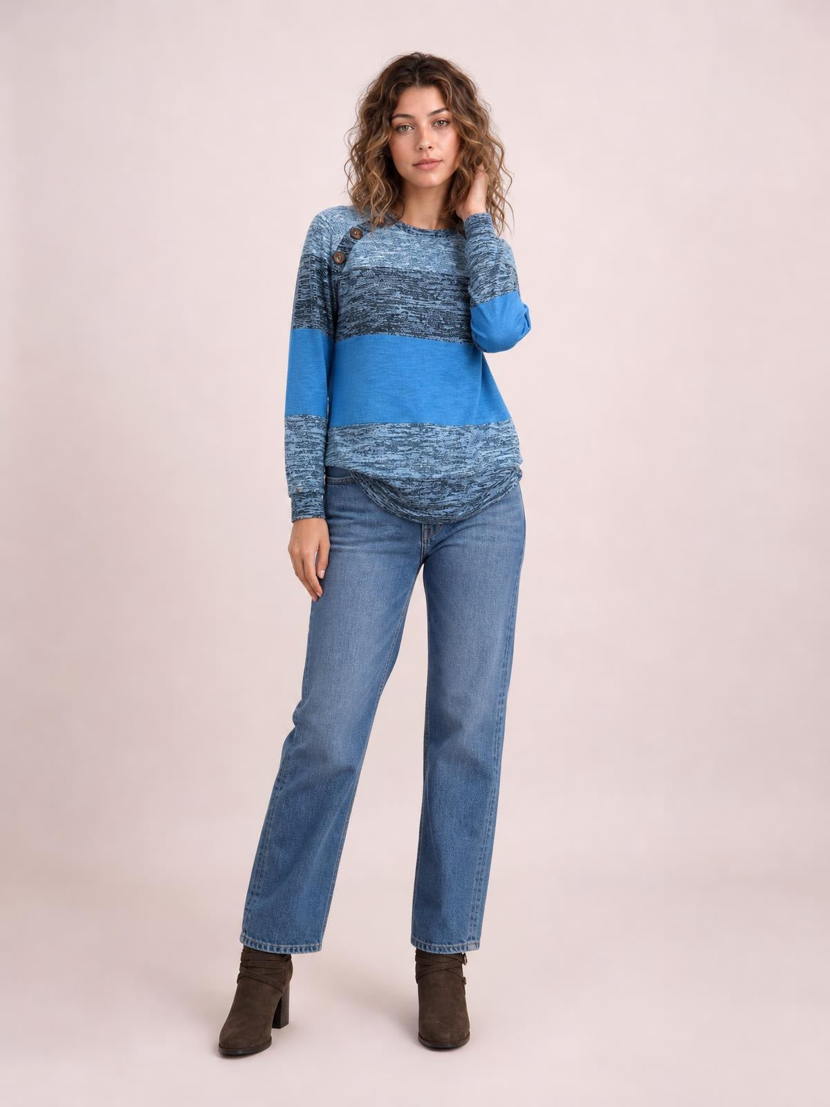 Model poses in a blue contrast round-neck long-sleeve T-shirt with decorative shoulder buttons, patterned panels, and a relaxed, curved hem.