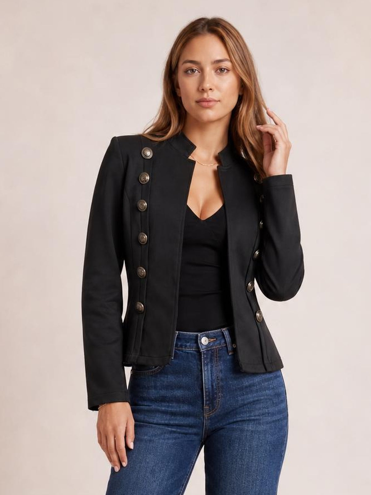 Model poses in a black open-front long-sleeve jacket with decorative buttons, structured shoulders, and a fitted silhouette over a black top and blue jeans.