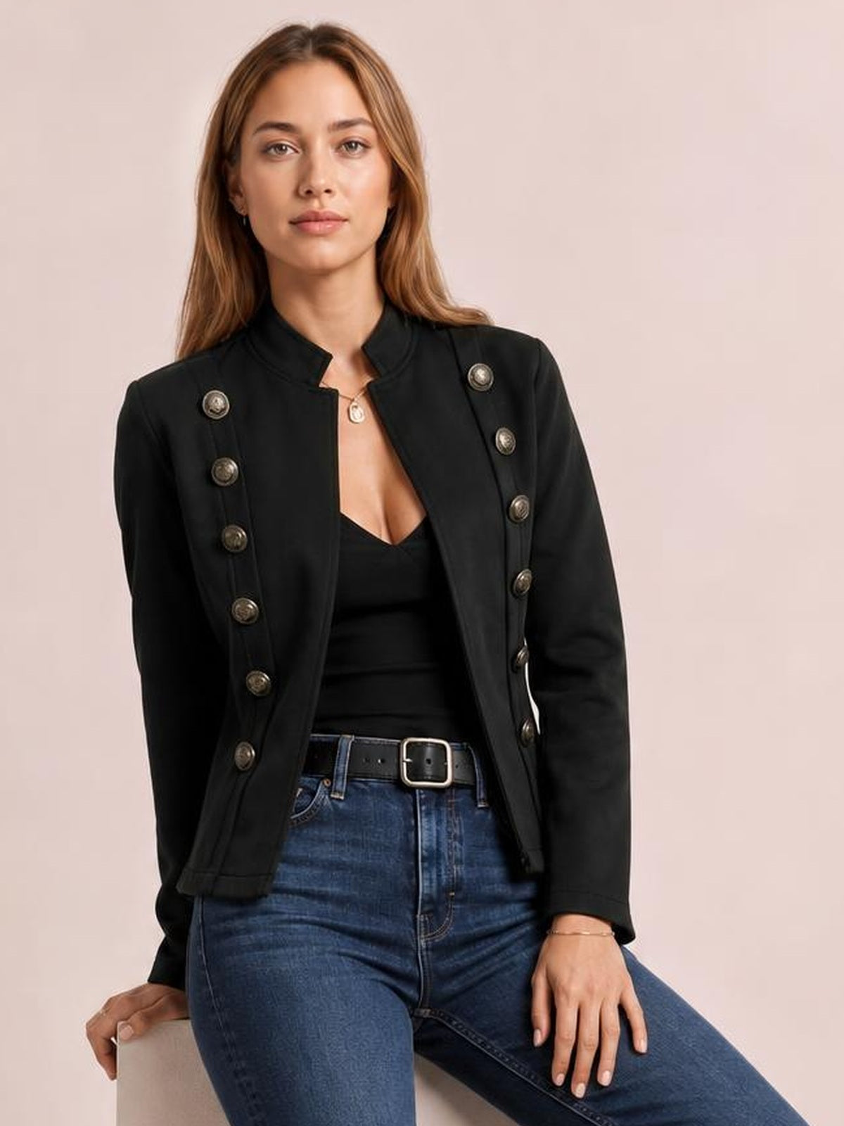 Model seated in a black open-front long sleeve jacket with a structured silhouette, stand collar, and rows of decorative buttons.