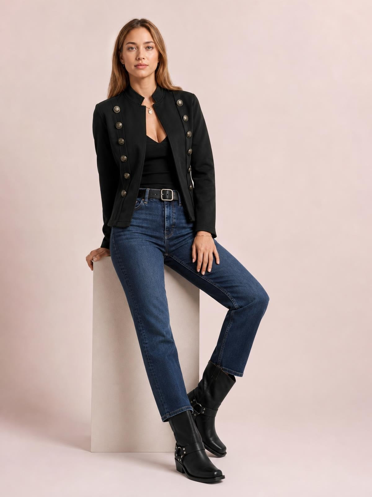Model seated in a black open-front long sleeve jacket with decorative buttons, structured shoulders, and a fitted silhouette over a black top and jeans.