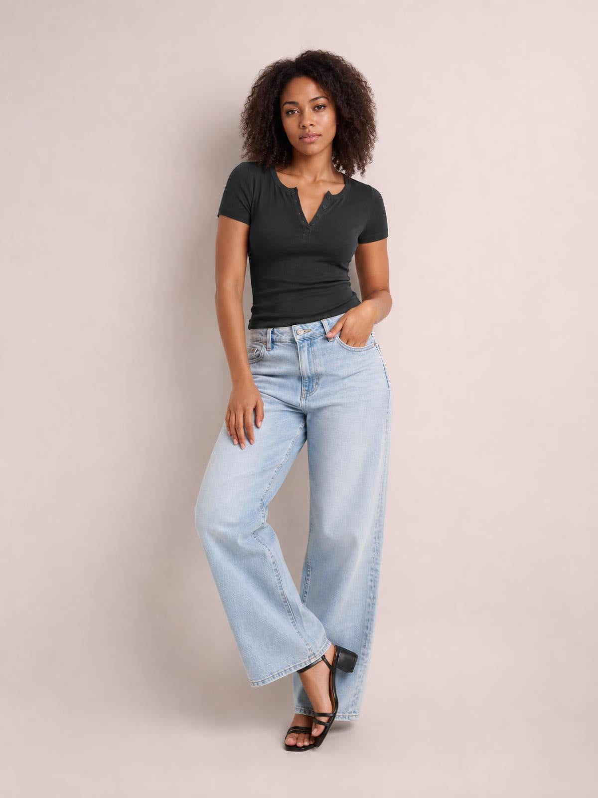Model wears a black button-detail short-sleeve T-shirt with a fitted silhouette and notched henley neckline, standing with one hand in her pocket and legs crossed.