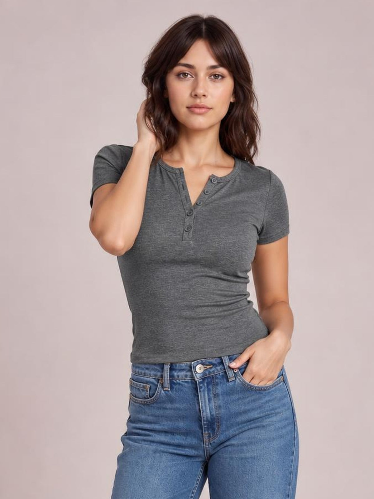 Model wears a charcoal gray short-sleeve T-shirt with a fitted silhouette, rounded neckline, and button detail, posing with one hand in her jeans pocket.