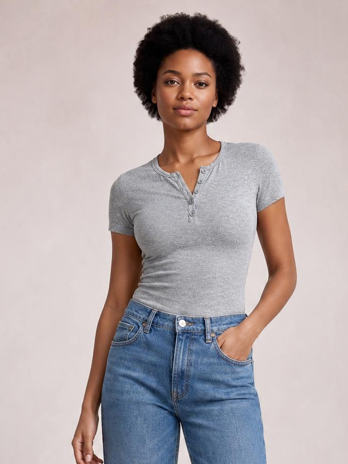 Model wearing a fitted gray short sleeve T-shirt with a round neckline, button-detail placket, and stretchy silhouette, posing with one hand in her jeans pocket.