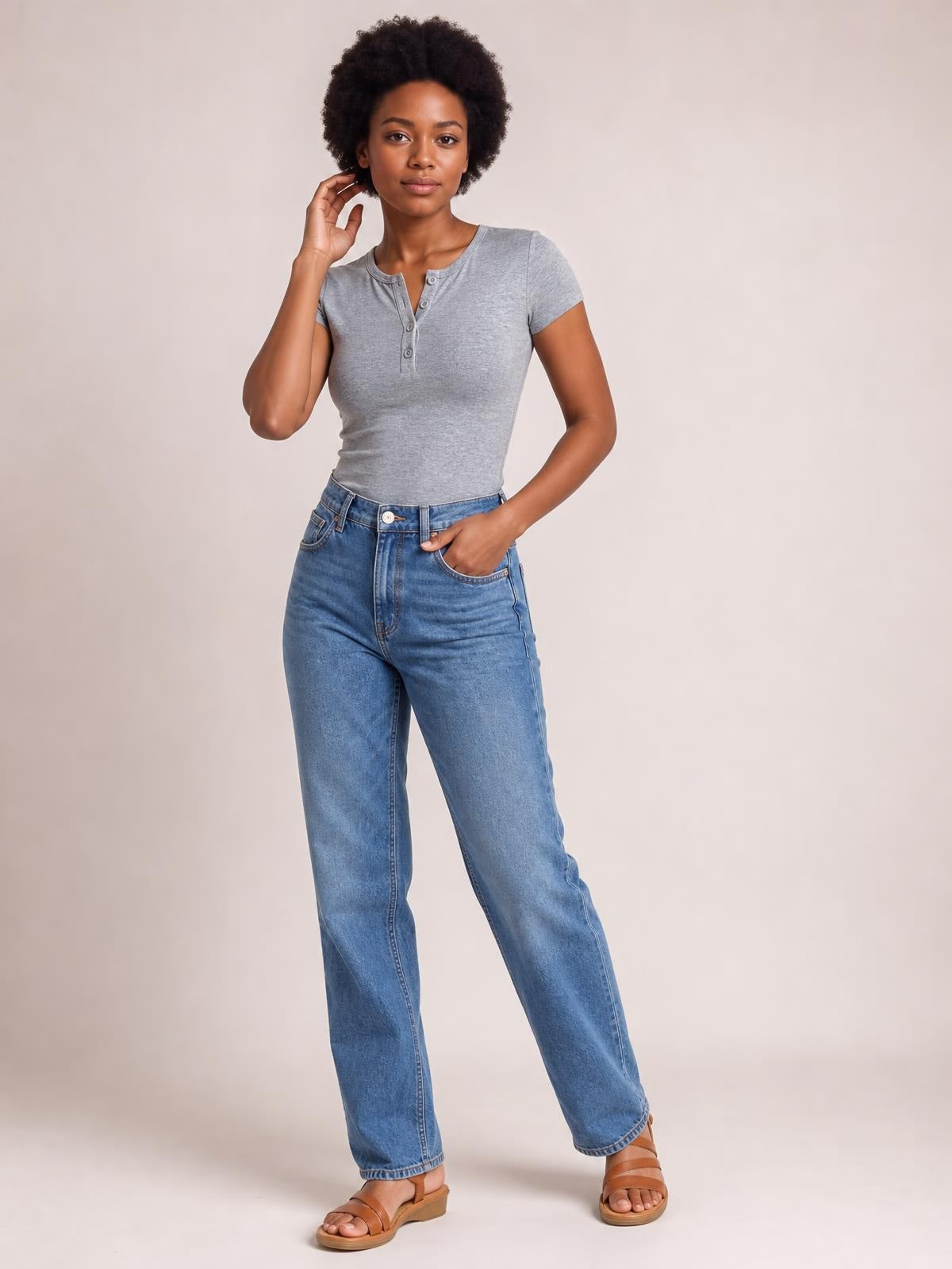 A woman poses in a fitted gray short-sleeve T-shirt with a buttoned split neckline, tucked into high-waisted jeans, one hand at her hair and the other in a pocket.