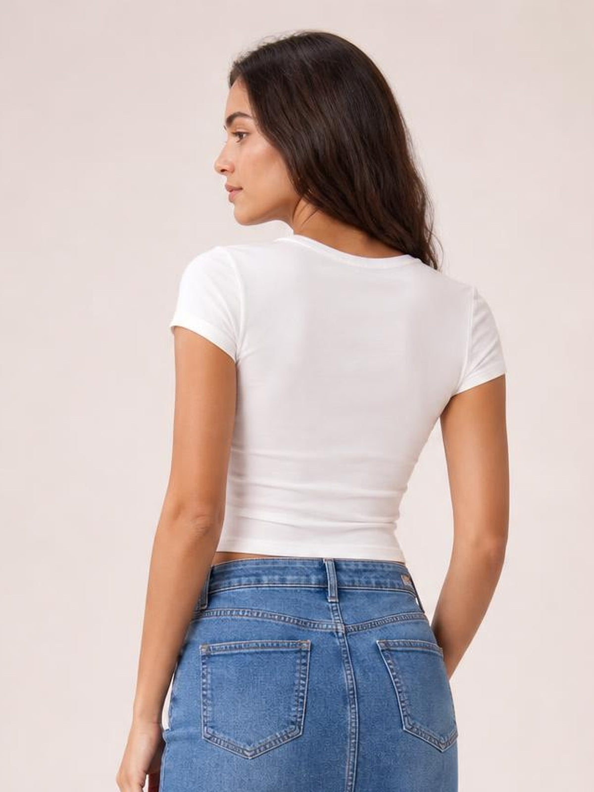 A model looks over her shoulder while wearing a white short sleeve T-shirt with a fitted silhouette, smooth opaque fabric, and a rounded neckline.