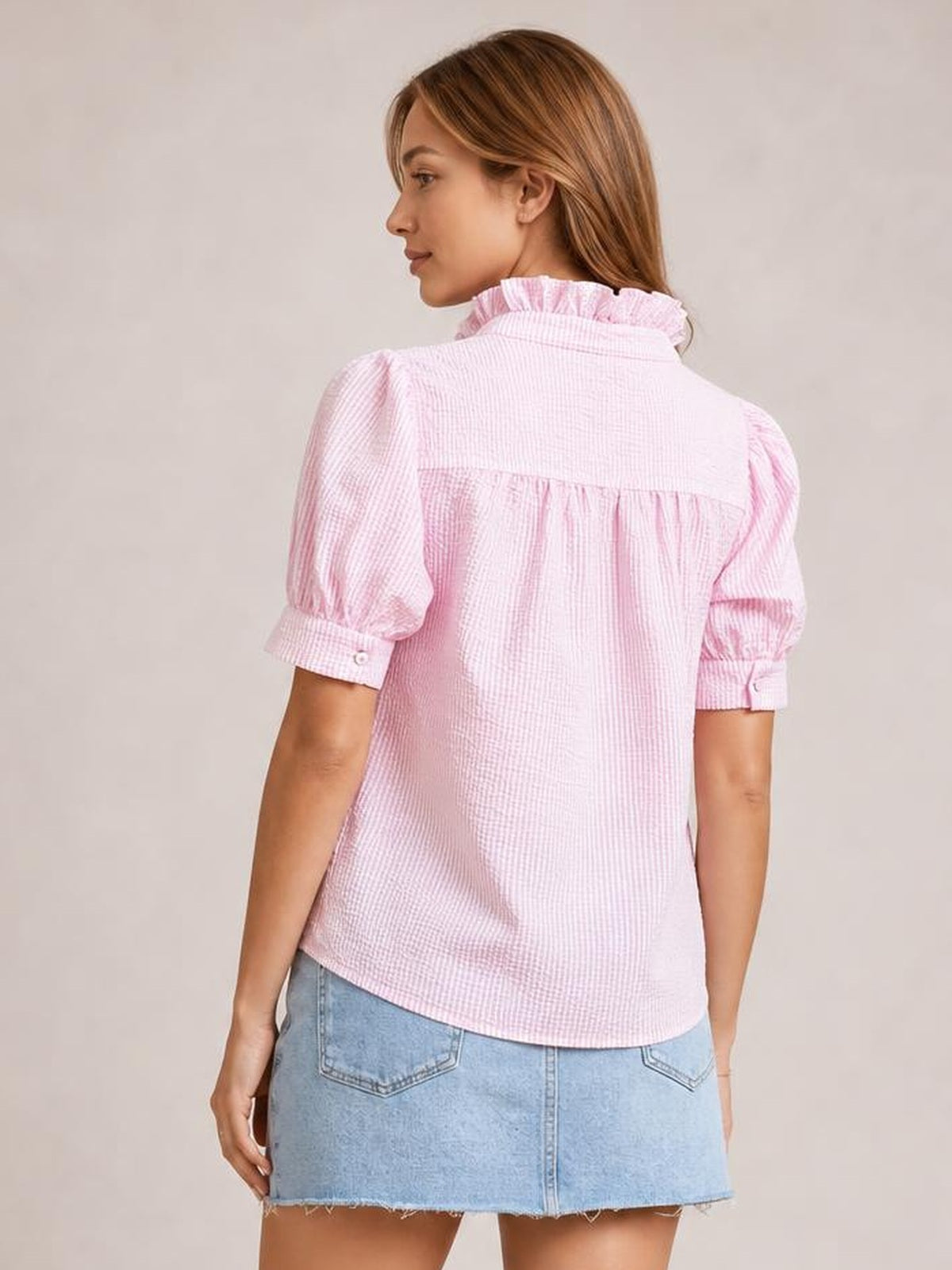 A model turns to show the back of a pink striped button-down shirt with a ruffled collar, puffed short sleeves, button cuffs, and a relaxed curved hem.