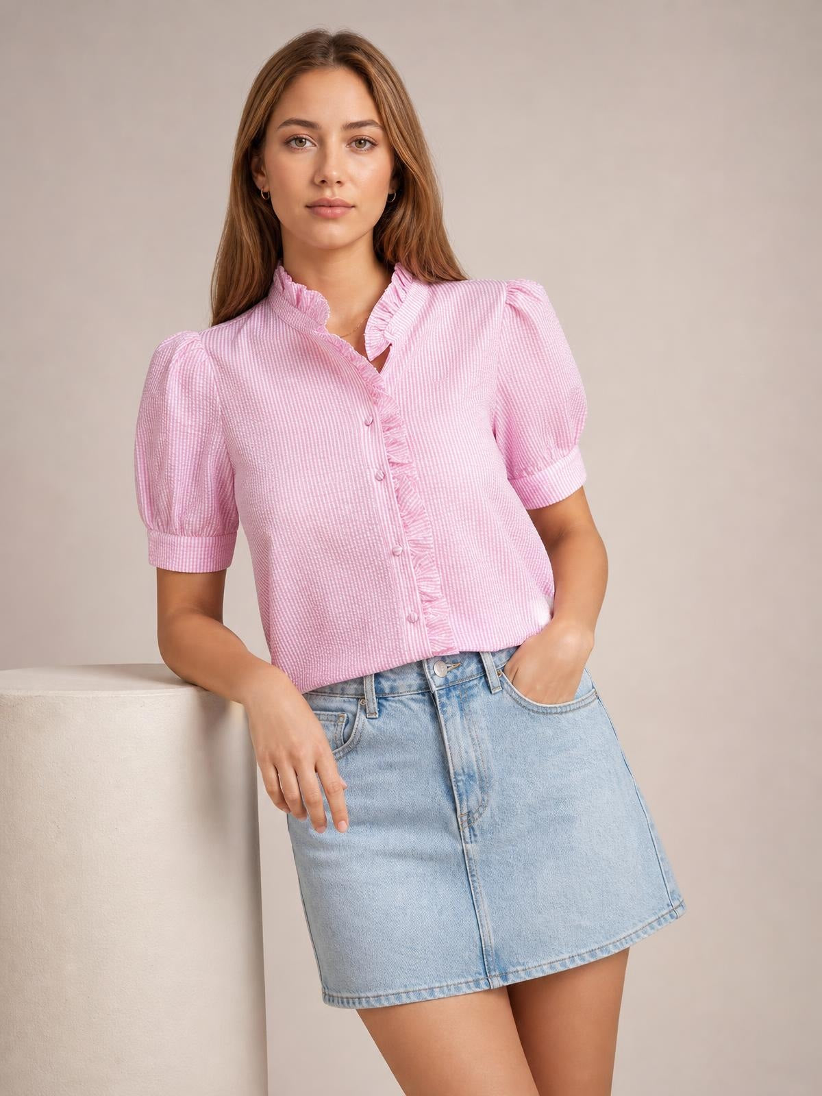 A model leans casually in a pink striped button-down shirt with short puff sleeves, ruffled trim, a stand collar, and a relaxed cropped silhouette.