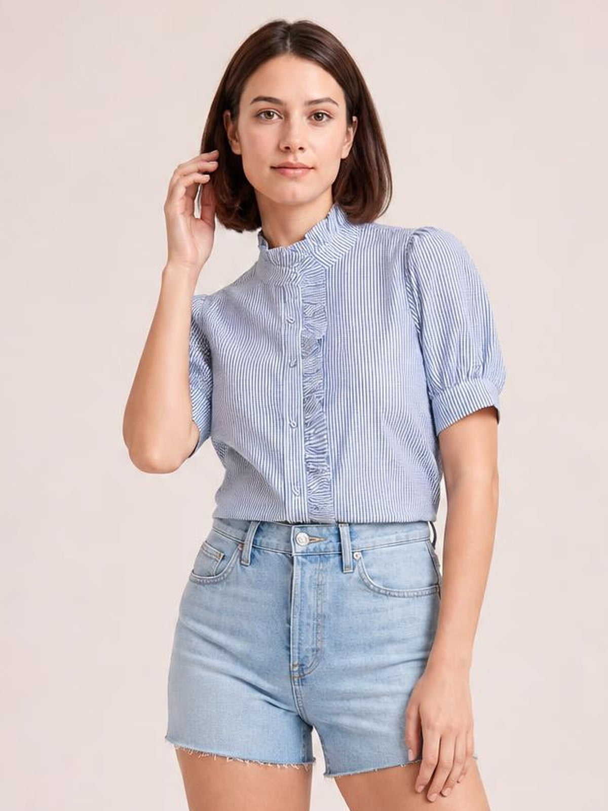 Model wearing a blue and white striped button-down short sleeve shirt with a high neckline, ruffled placket, and relaxed cropped silhouette, touching her hair.