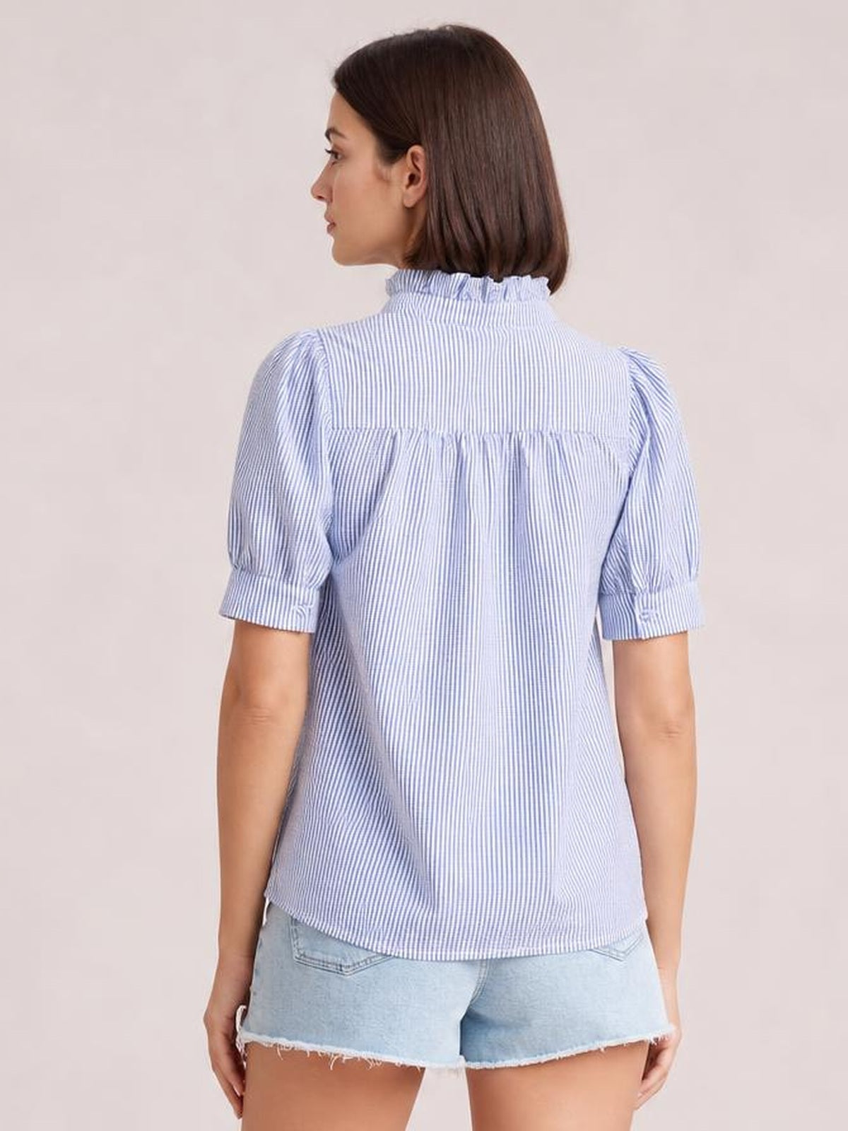 Model shown from behind wearing a light blue striped short sleeve shirt with a button-down front, ruffled collar, gathered back, and relaxed silhouette.