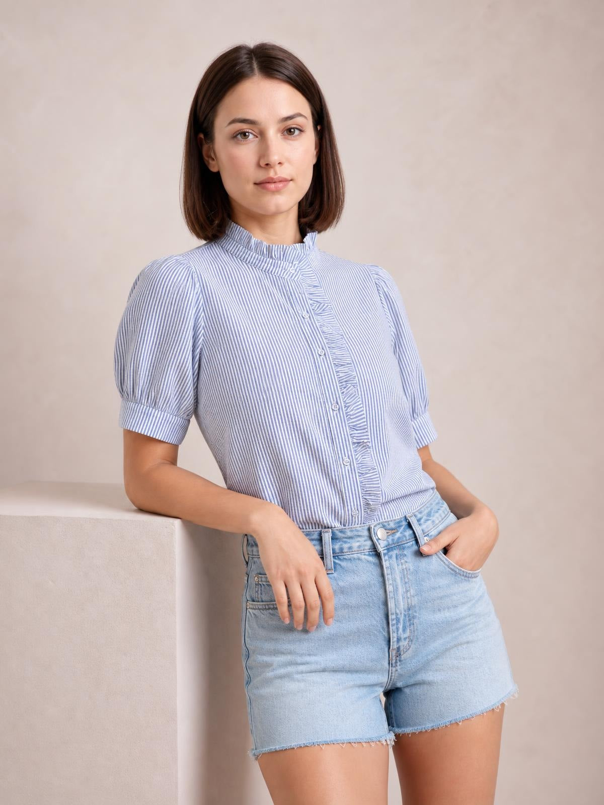A model leans casually in a light blue and white striped button-down shirt with short puff sleeves, a ruffled high collar, and buttoned front.